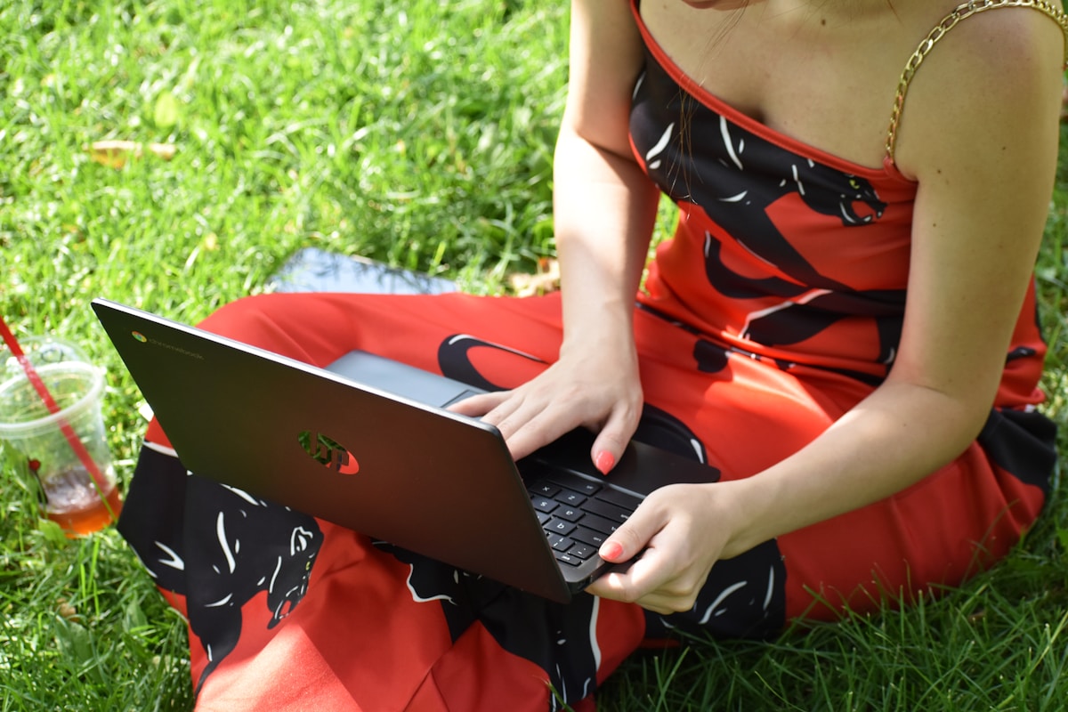 Femme assise sur l'herbe, utilisant un ordinateur portable rouge et noir.
